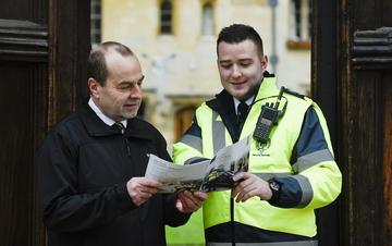 Photo of a male security officer wearing a high-vis jacket looking at a brochure with a male college porter